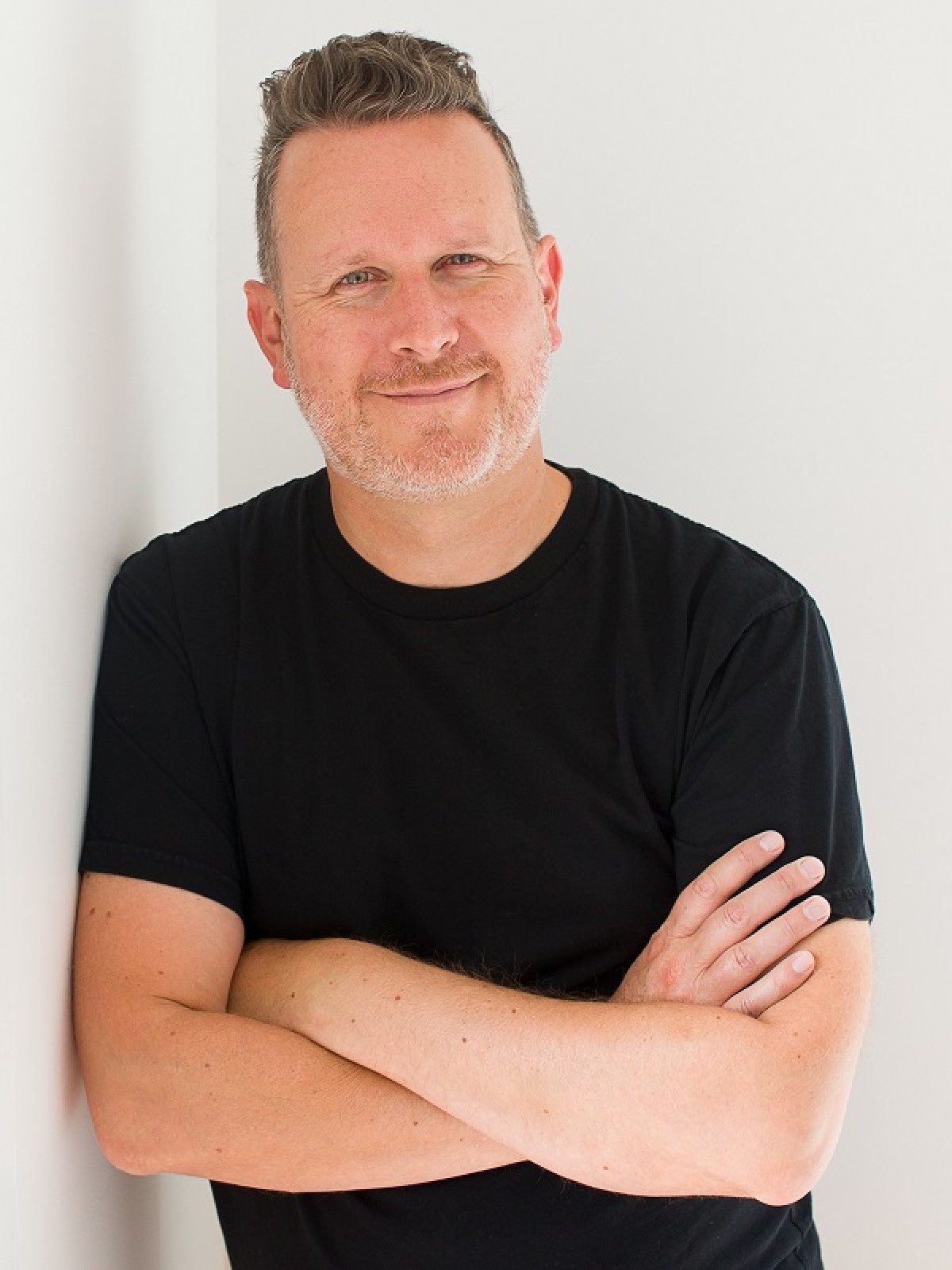 Man in a black T-shirt, friendly smile, leans against a wall to his right.